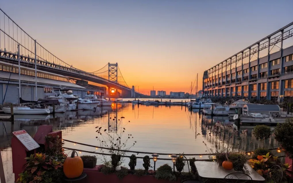 Golden hour at Pier 5 Marina, looking across still water and moored boats toward the Ben Franklin Bridge, with a planted marina deck in the foreground